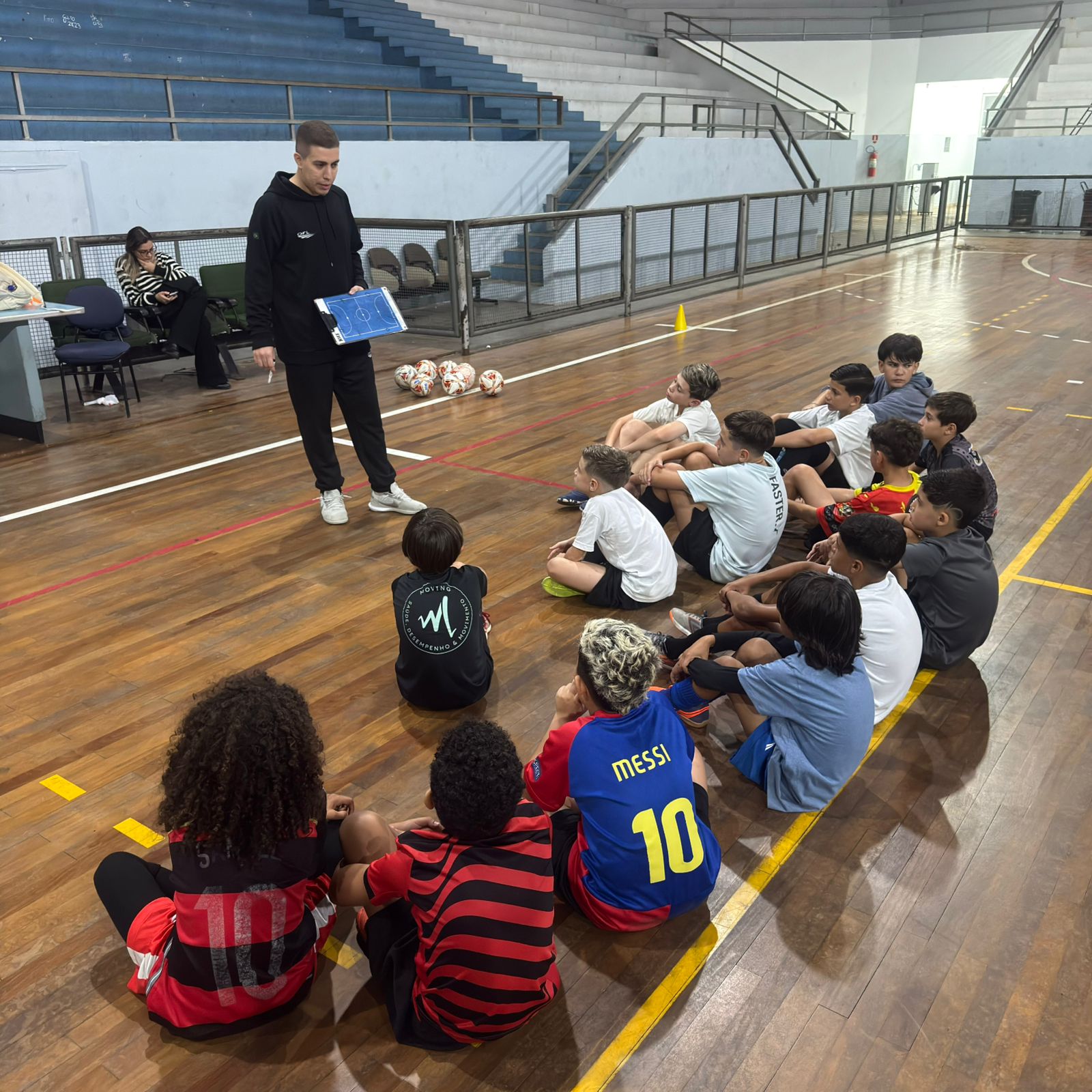 Treinador Fernando orientando crianças sentadas em roda na quadra da FEG